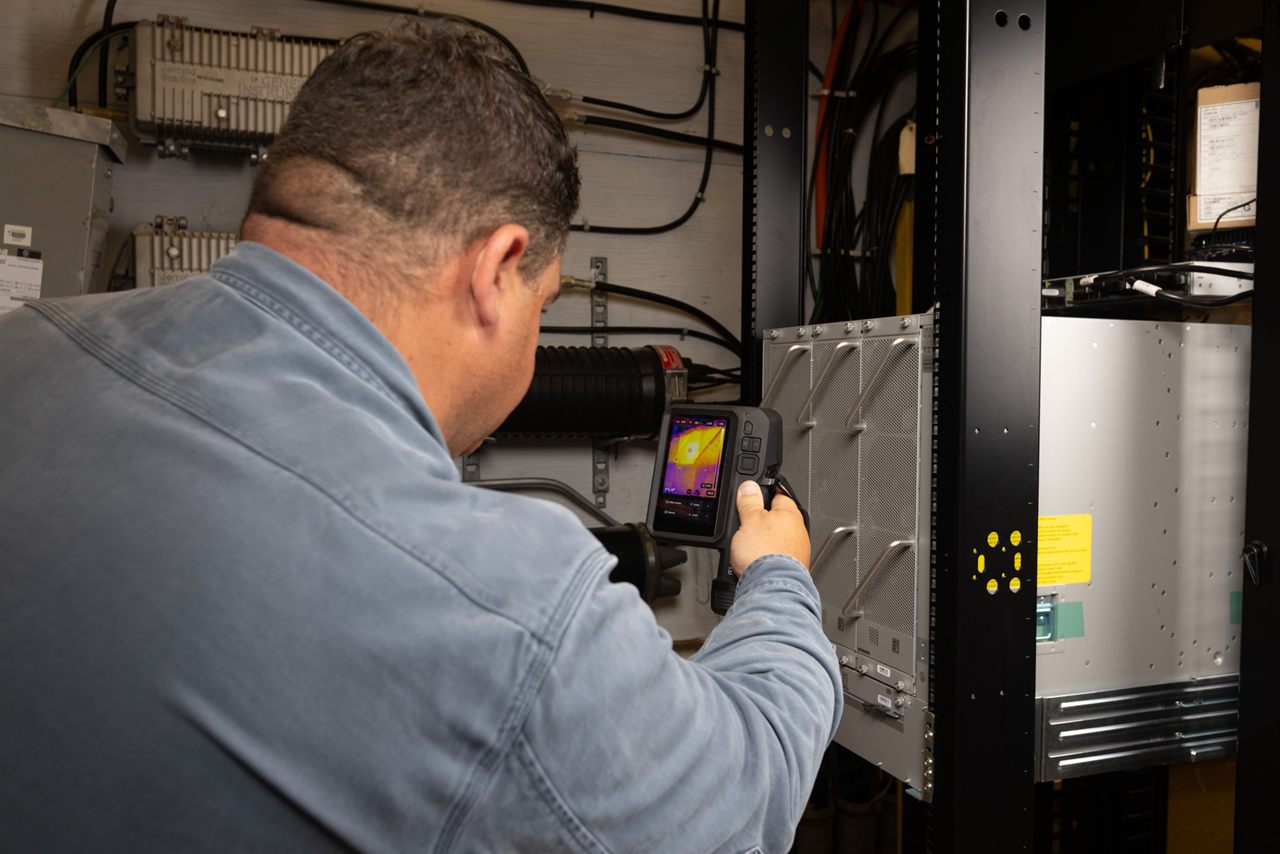 Technician thermally scanning a data-centre server rack with a FLIR camera, a hotspot visible on the screen.