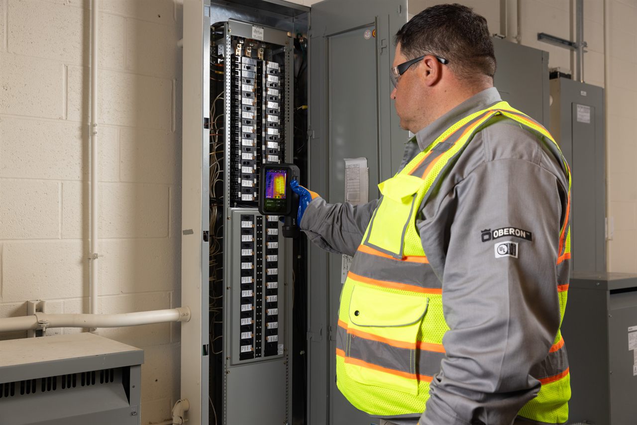 A hi-vis electrician scans an open electrical switchboard with a handheld FLIR thermal camera; a live thermal image is visible on the camera screen.